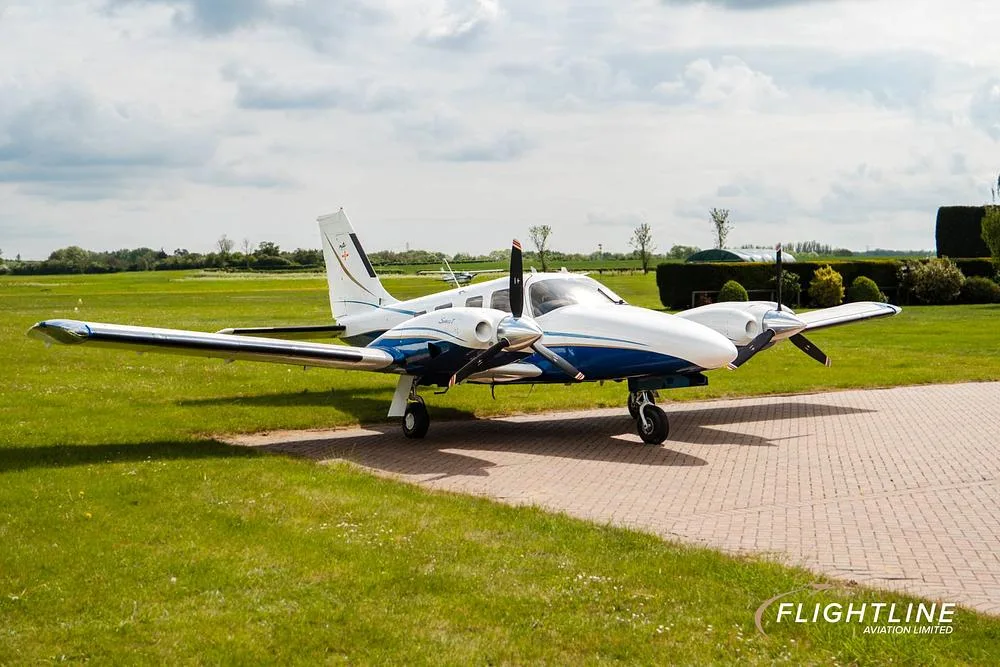 A front-side view of a white and blue twin-engine Piper Seneca aircraft parked on a tarmac.