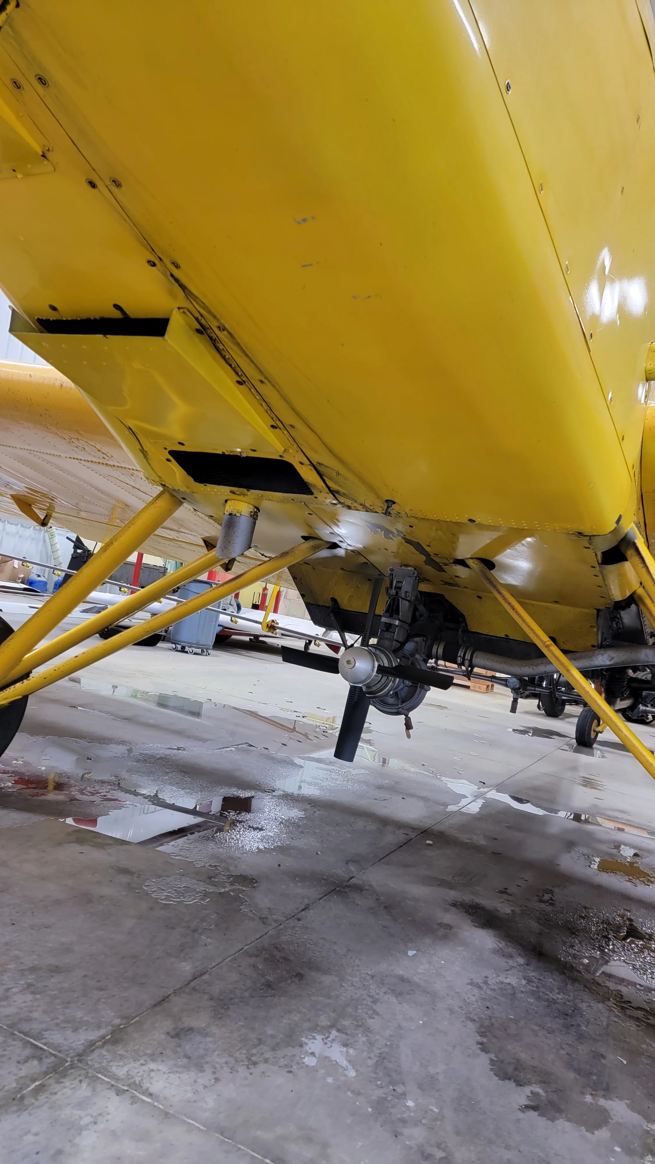 A low-angle view of the yellow aircraft's underside, showing the landing gear and spray nozzles in a hangar.
