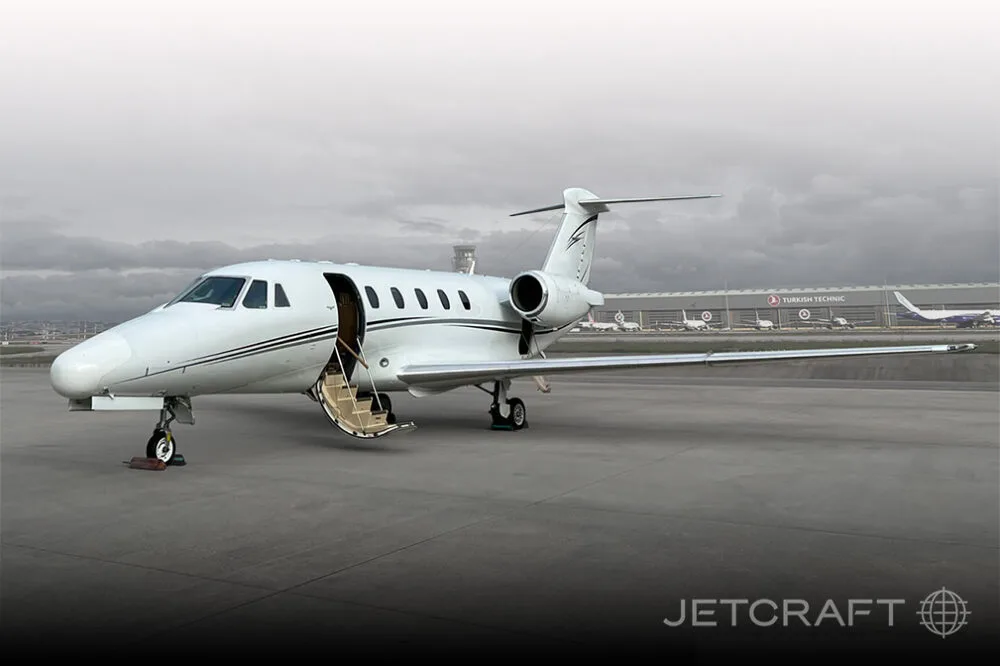 A white Cessna Citation business jet with its door open and stairs extended, parked on an airport tarmac under a cloudy sky.