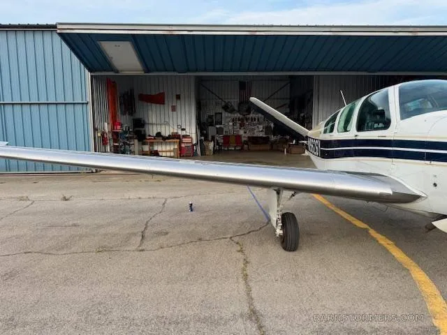 A side view of the wing and fuselage of a white and blue Beechcraft Bonanza, parked next to an open hangar with tools visible inside.
