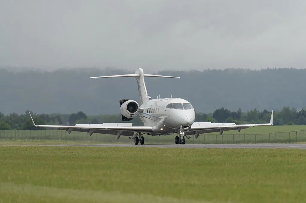 A white business jet with a blue stripe on a runway, viewed from the front, with green grass and hills in the background under a cloudy sky.