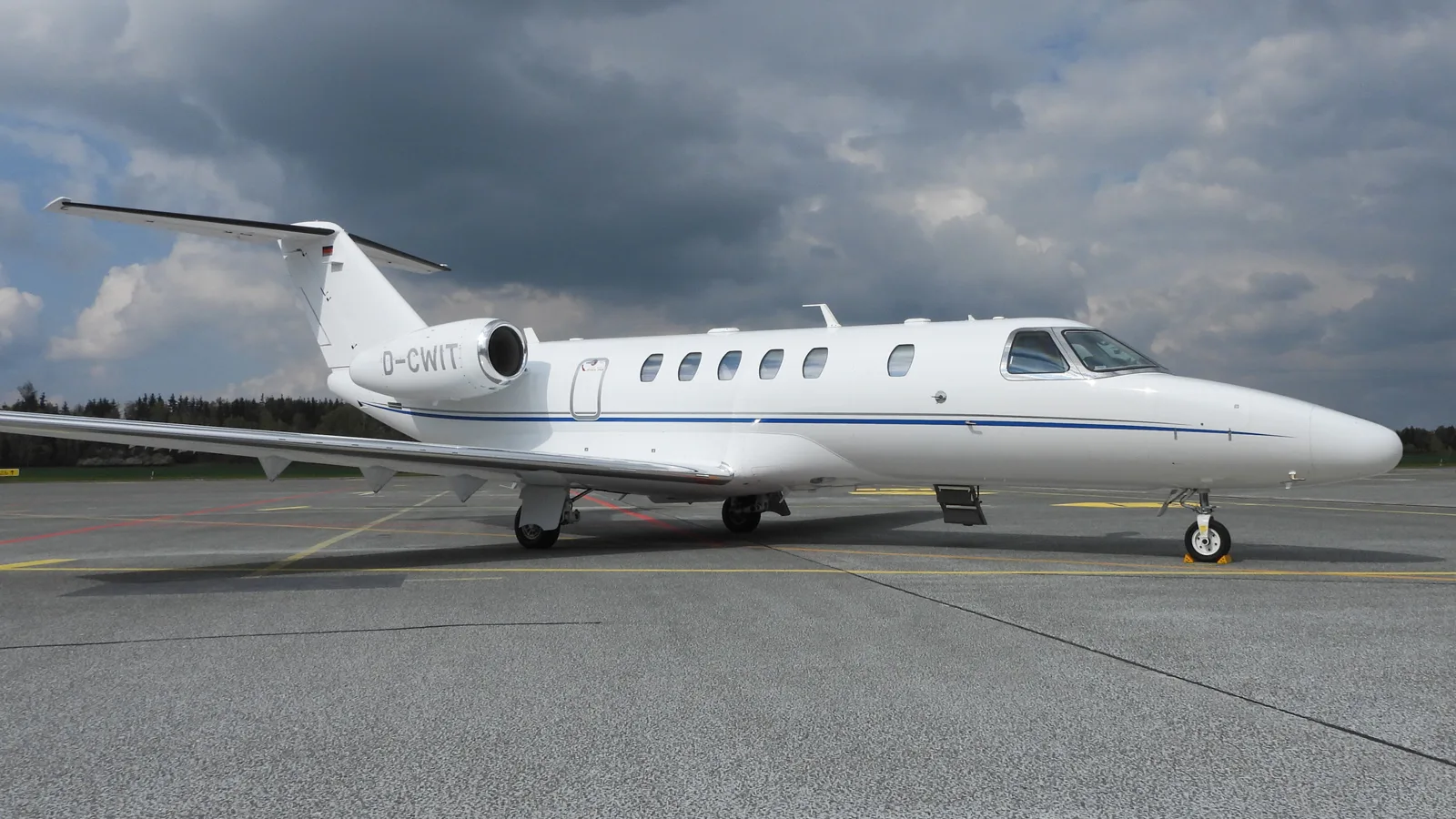 A white Cessna Citation CJ4 business jet with blue stripes, registration D-CWIT, parked on the tarmac under a cloudy sky.