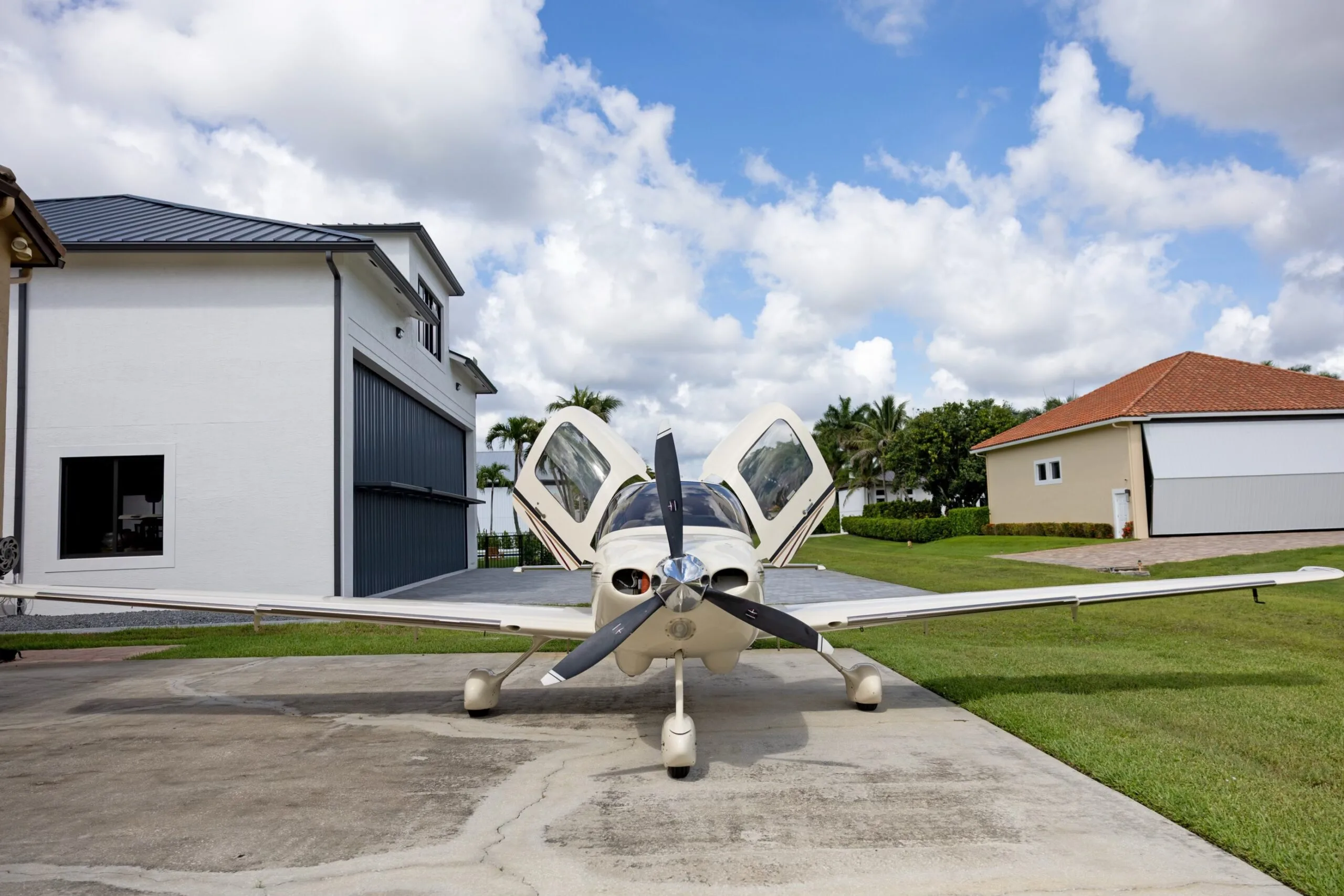 A front view of a white Cirrus SR22 aircraft with its gull-wing doors open, parked on a concrete pad between two buildings.