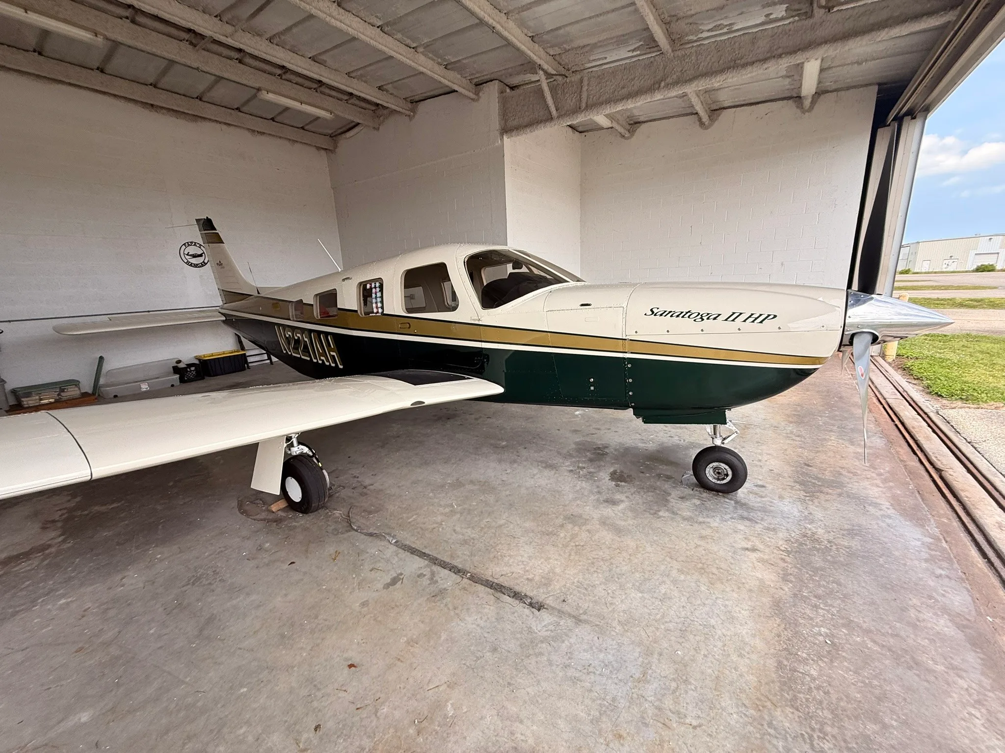 Side view of a Piper Saratoga II HP, N221AH, in a white, gold, and dark green paint scheme, parked inside a hangar.