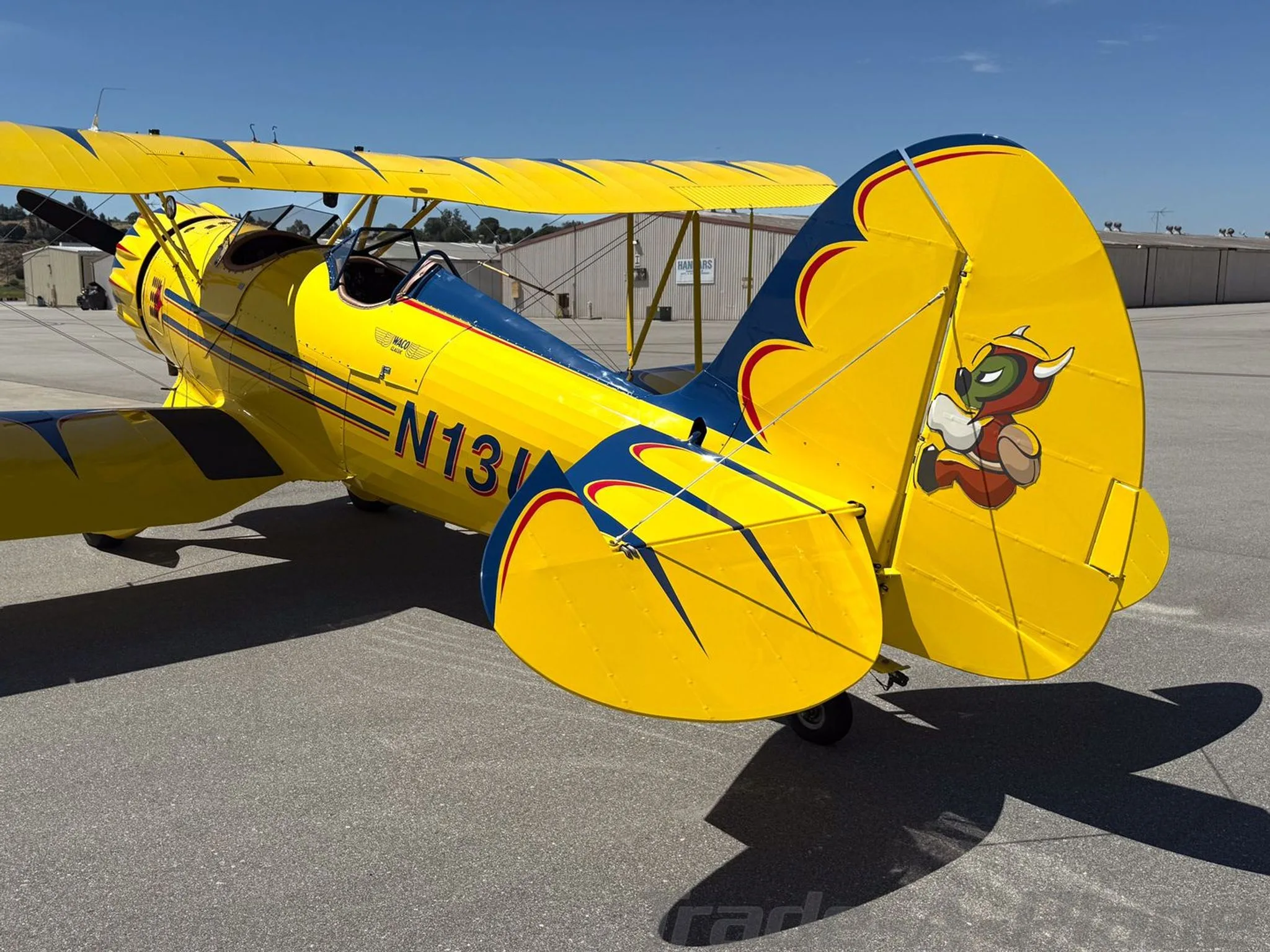 A rear three-quarter view of the yellow and blue biplane N13UP, showing the tail with a cartoon devil.