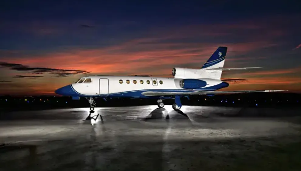 A white and blue Dassault Falcon business jet with a tail logo, parked on a wet tarmac at sunset.
