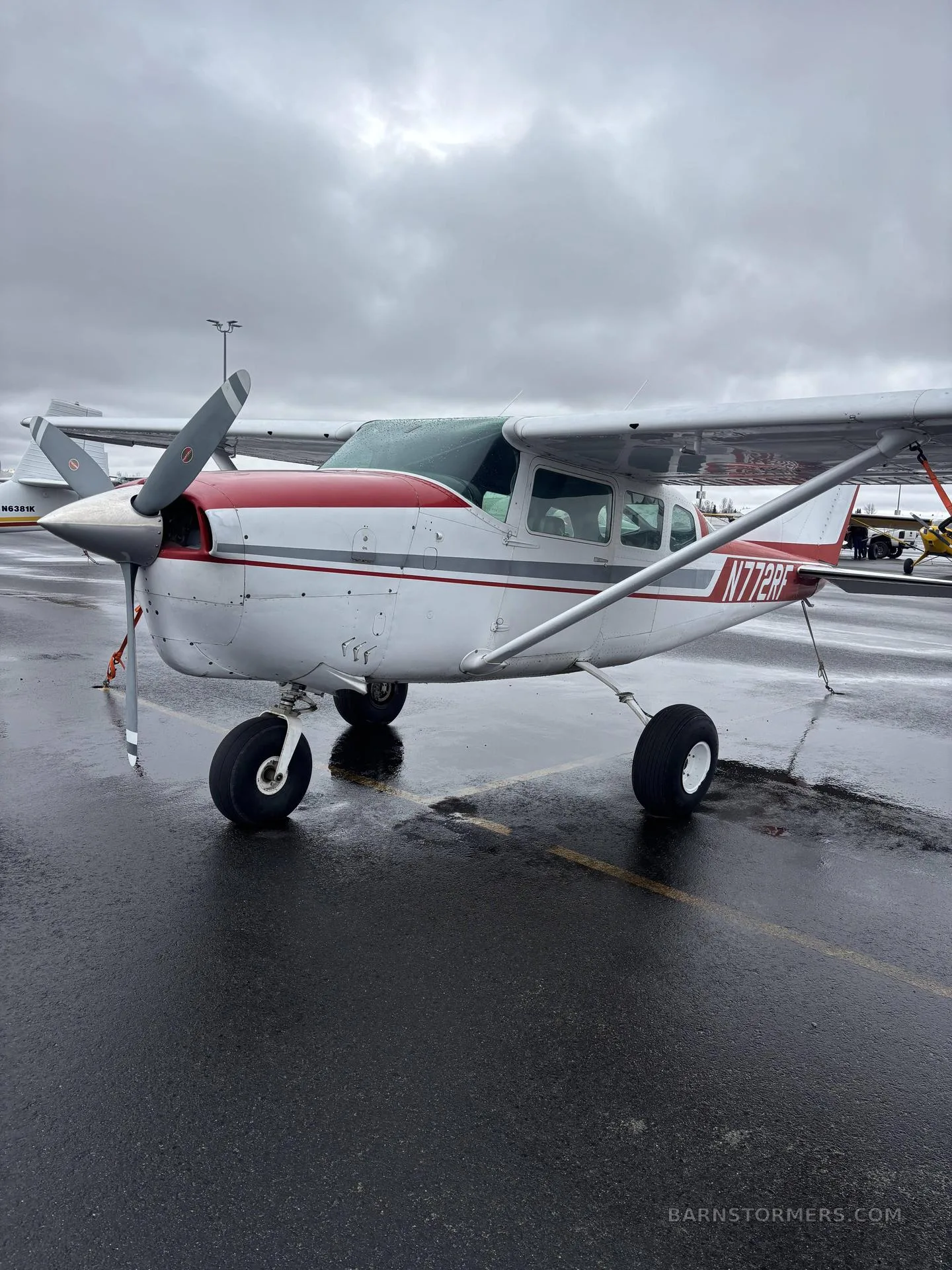 A white and red single-engine Cessna aircraft, registration N772RF, parked on a wet tarmac under a cloudy sky.