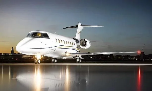A white business jet with blue and gold stripes parked on an airfield at dusk, reflecting lights on the wet tarmac.