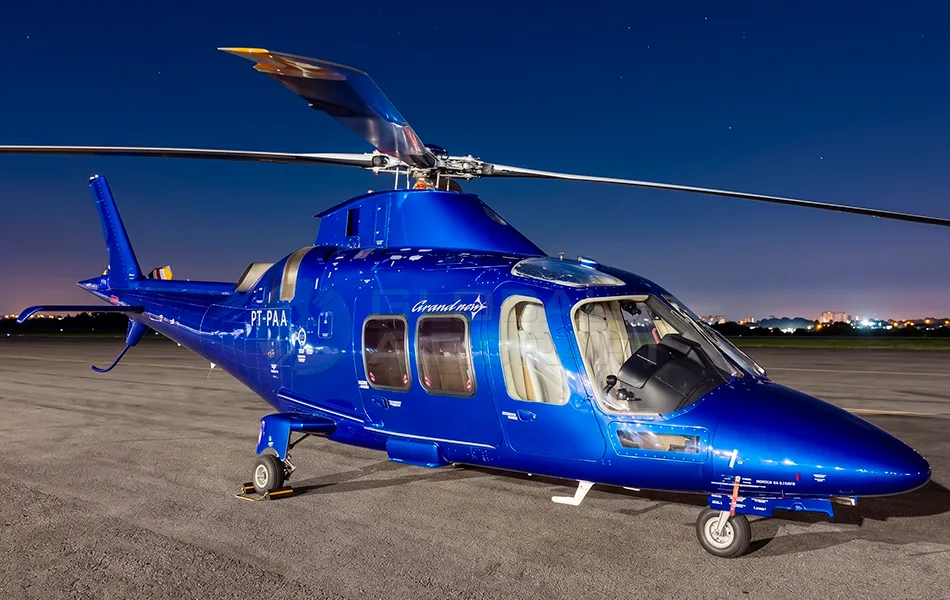 A shiny blue AgustaWestland AW109 GrandNew helicopter with registration PT-PAA is parked on an tarmac at night, with city lights in the background.