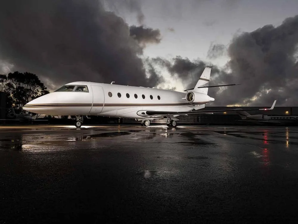 A white business jet with gold and black stripes parked on a wet tarmac under a dramatic cloudy sky at dusk.