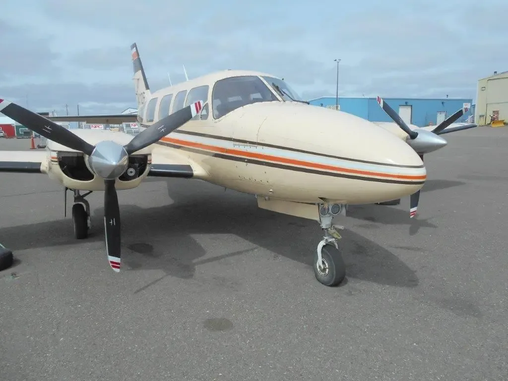 A front-quarter view of the beige twin-engine Piper Navajo aircraft, N4112E, with orange and black stripes, parked on an asphalt tarmac.