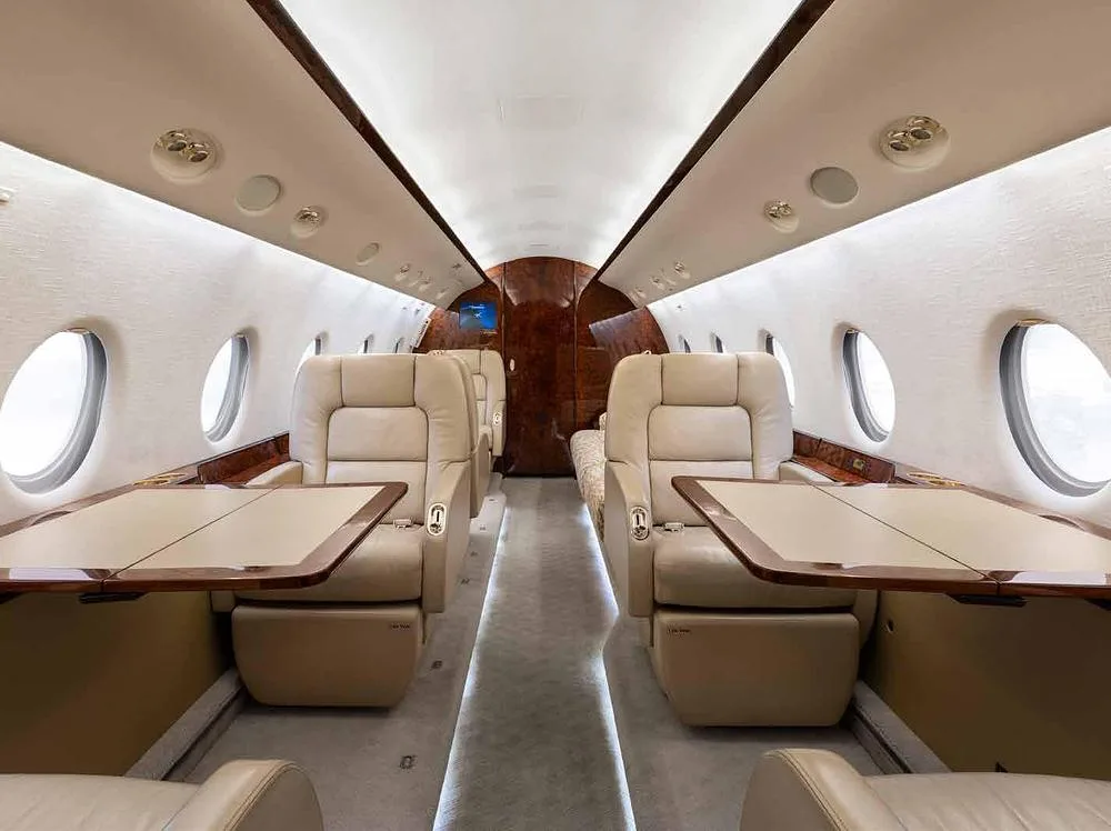 The luxurious interior cabin of a private jet with beige leather seats, deployed tables, wood paneling, and large oval windows.