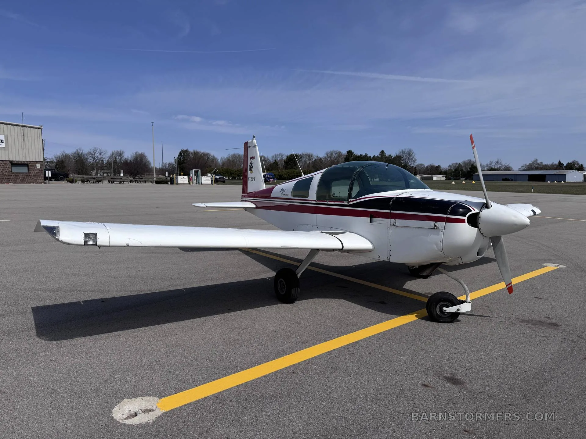 A white and maroon Grumman American AA-5 Traveler aircraft is parked on an asphalt tarmac under a clear blue sky.