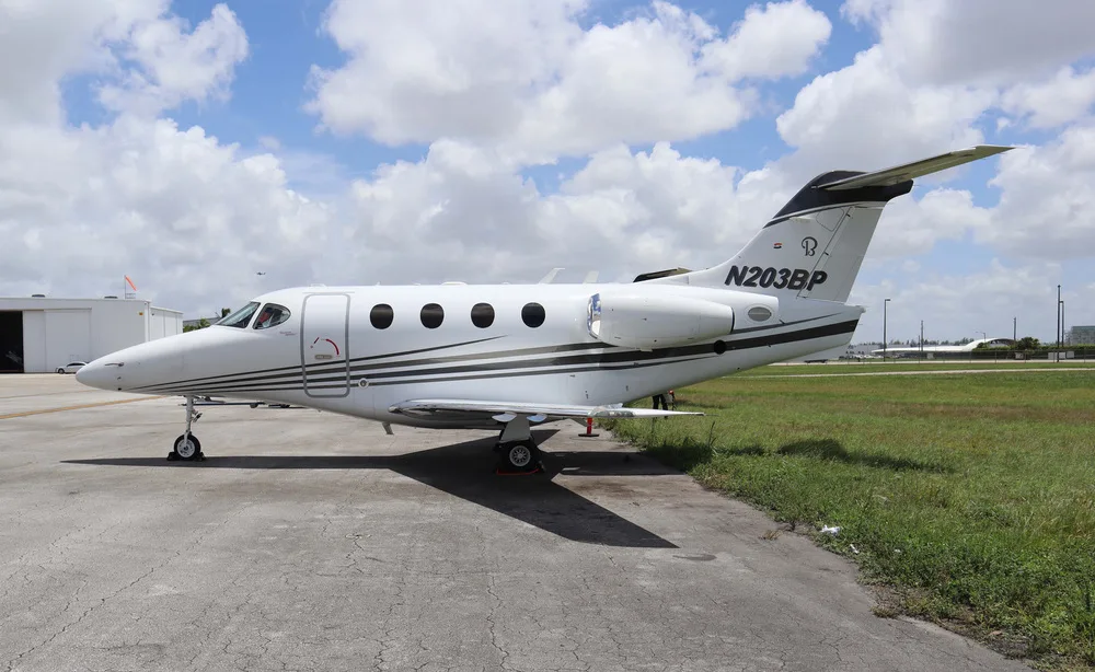A white Embraer Phenom 100 business jet with black and gold stripes on the fuselage and tail, parked on an asphalt ramp under a partly cloudy sky.