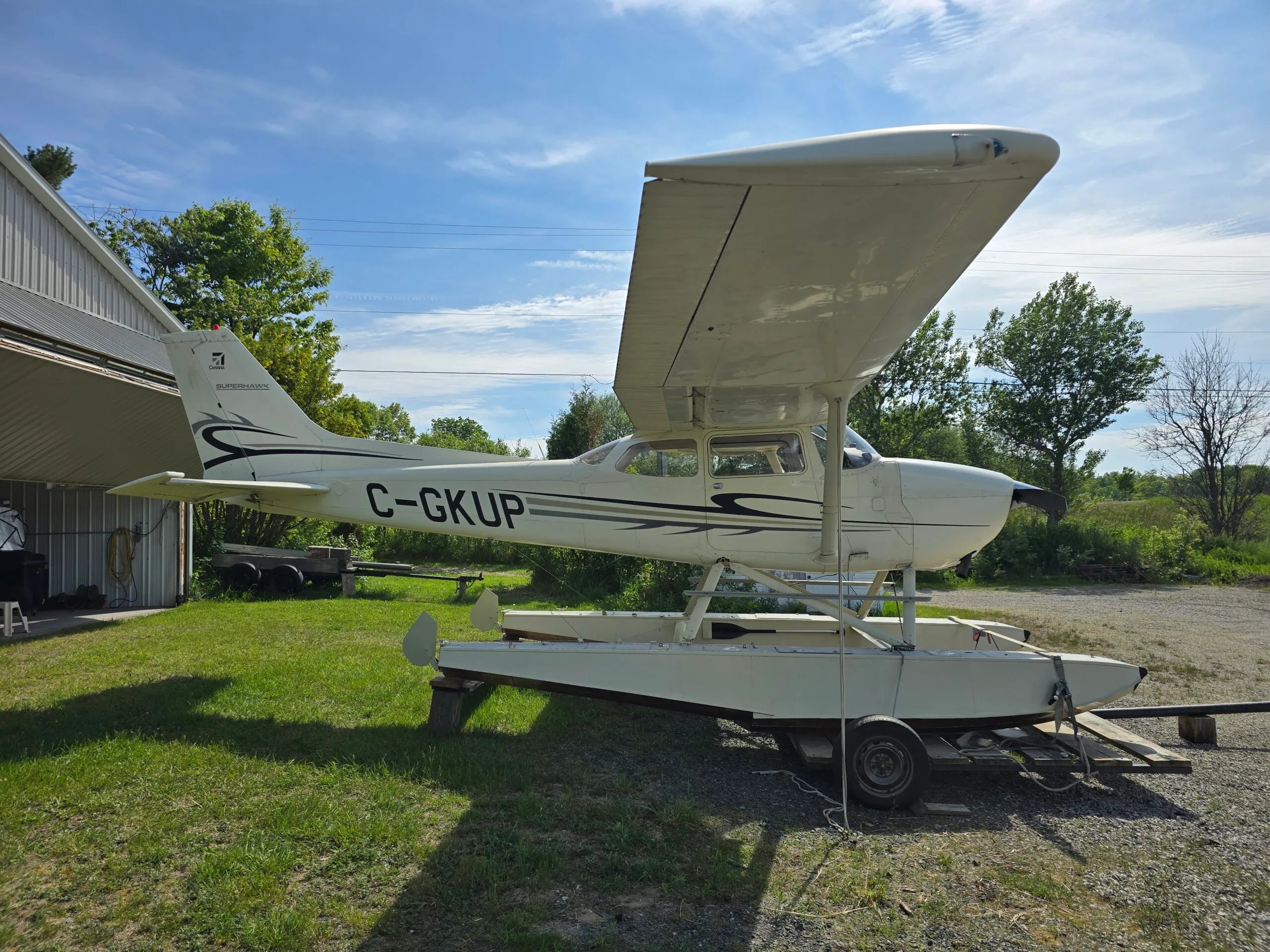 A white Cessna Superhawk floatplane with black and grey stripes is parked on a trailer next to a hangar on a sunny day.