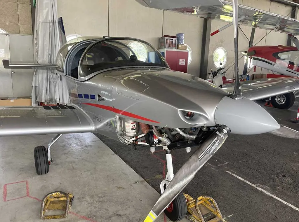 Front view of a silver Sonaca 200 aircraft with red accents, engine cowling open, parked in a hangar.