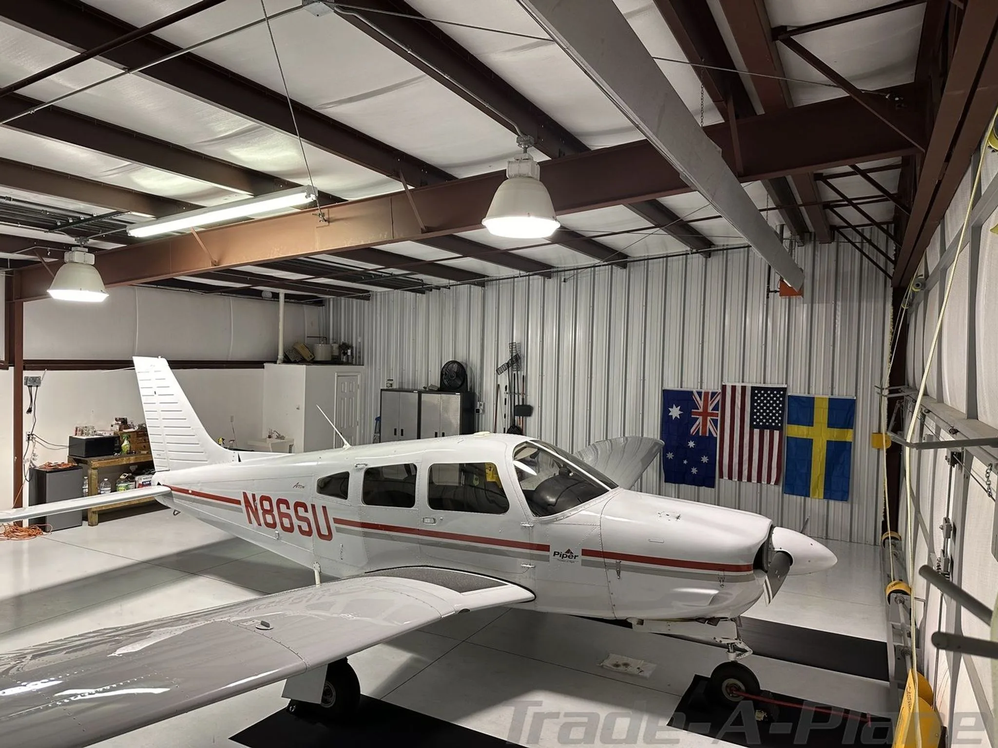 A white Piper aircraft with red stripes and tail number N86SU is parked inside a hangar with flags hanging on the back wall.