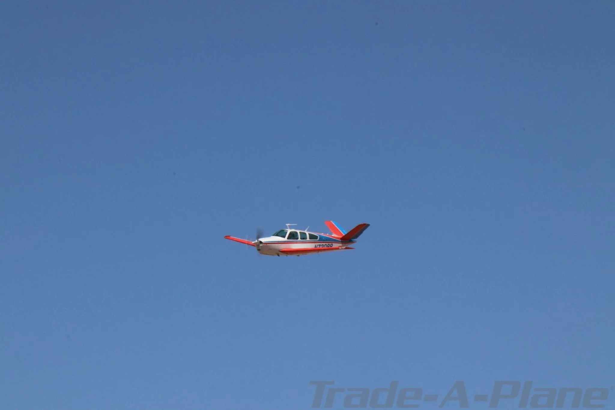A white, red, and blue Beechcraft Bonanza N7208R is flying in a clear blue sky.