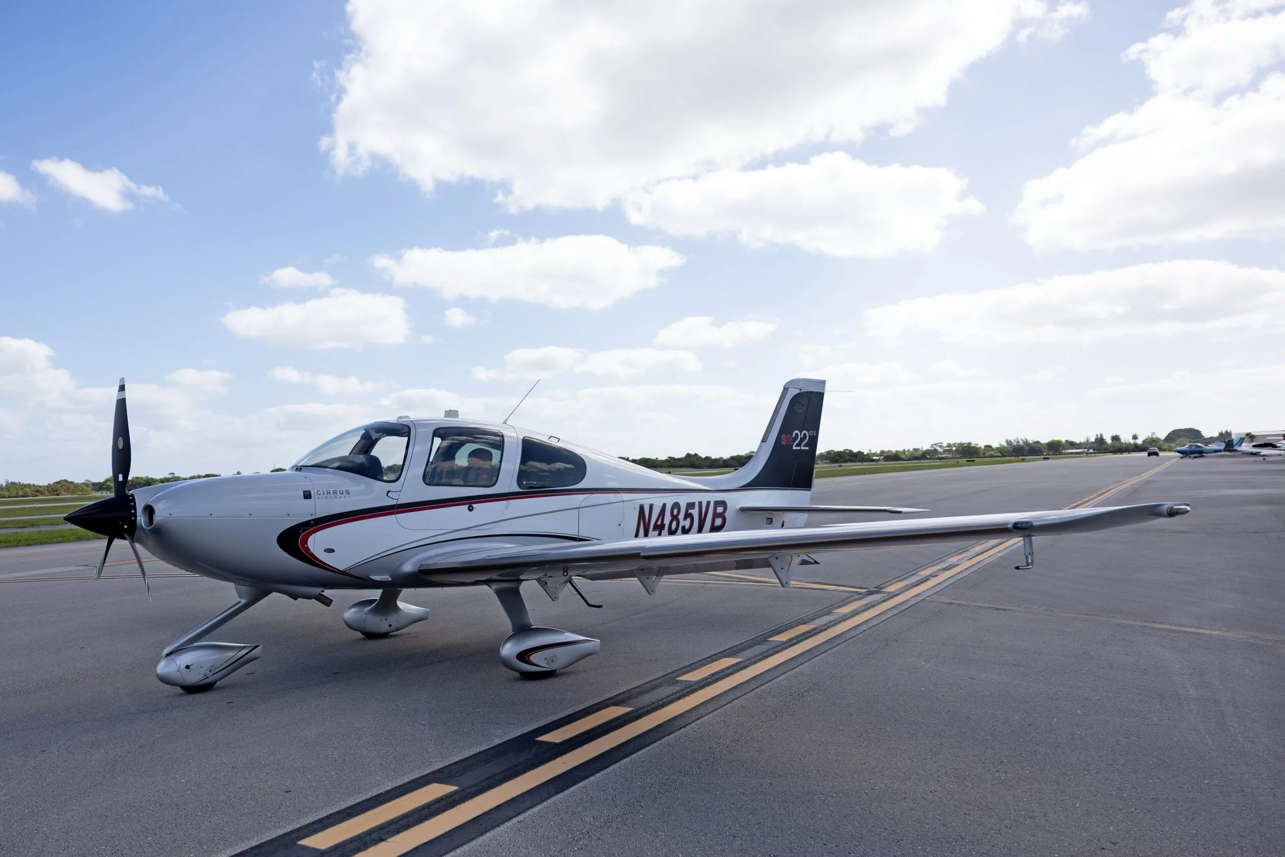 A side-quarter view of the silver Cirrus SR22 GTS aircraft N485VB, parked on an airfield under a bright, partly cloudy sky.