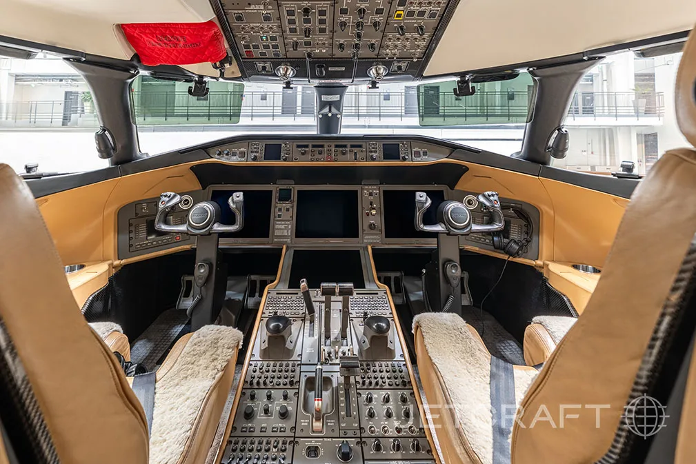 The cockpit of a private jet features a modern glass panel display, multiple control panels, and two pilot seats with sheepskin covers.