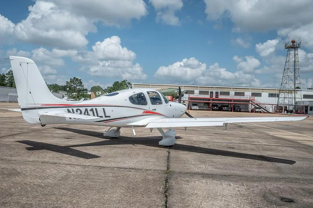 A white Cirrus SR22 aircraft, registration N241LL, parked on an airfield with a control tower and other aircraft in the background.