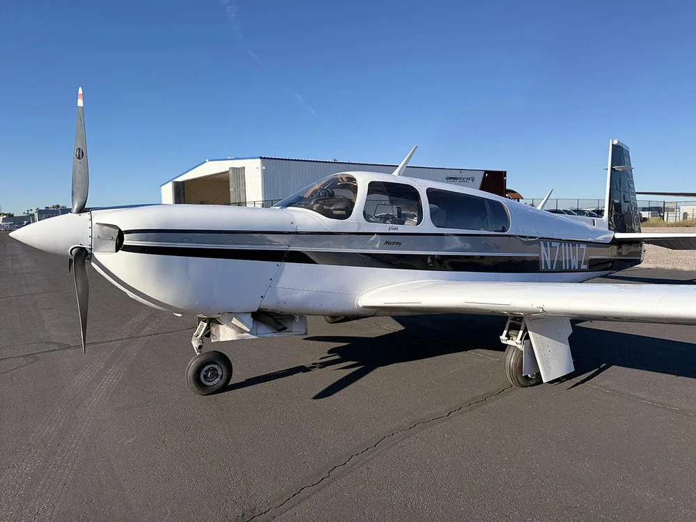 A front-left view of a white Mooney aircraft with black and grey stripes, tail number N711WZ, parked on an airfield.