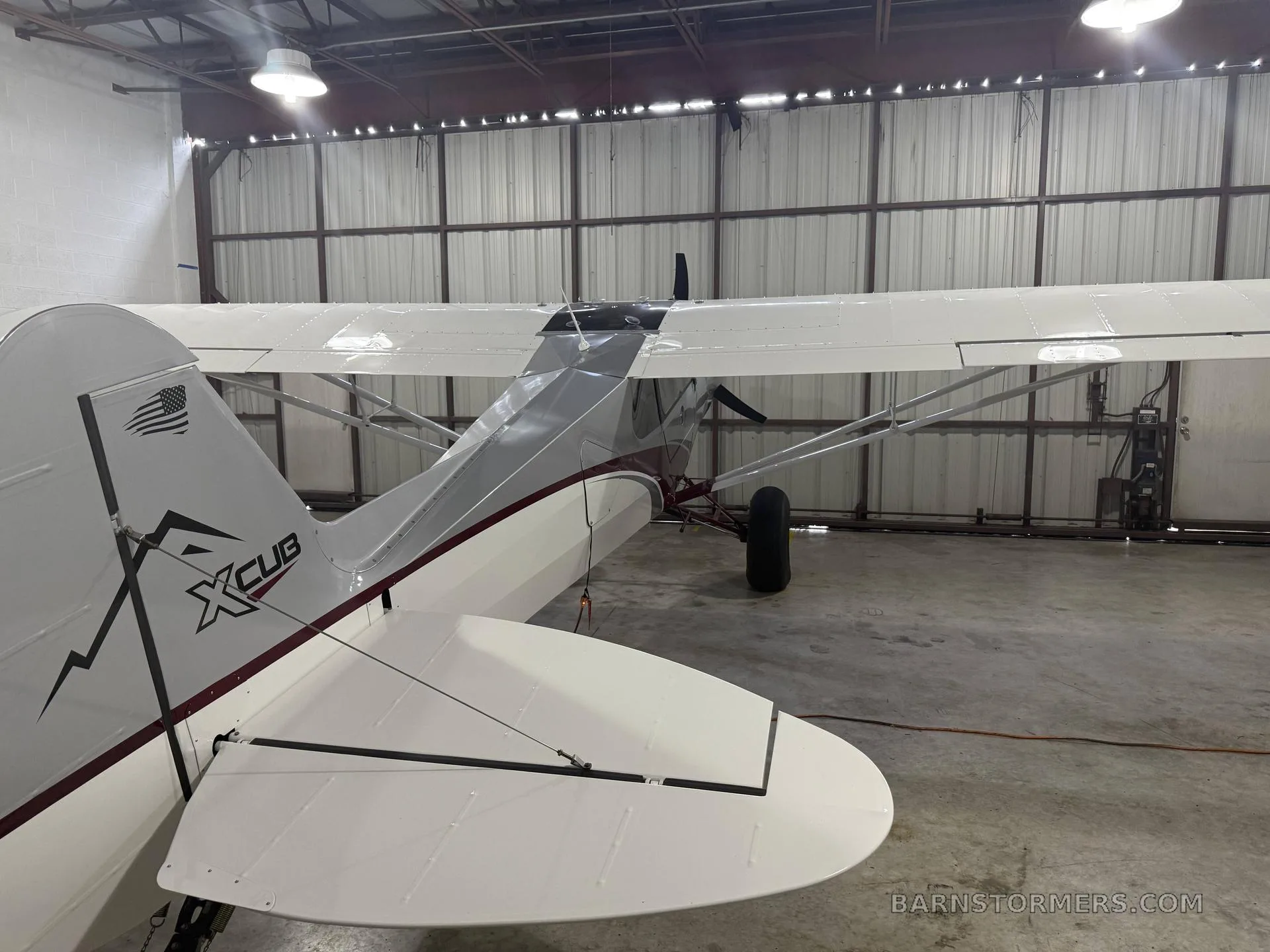 The rear view of a silver and red X-Cub aircraft with an American flag decal on the tail, parked in a hangar.