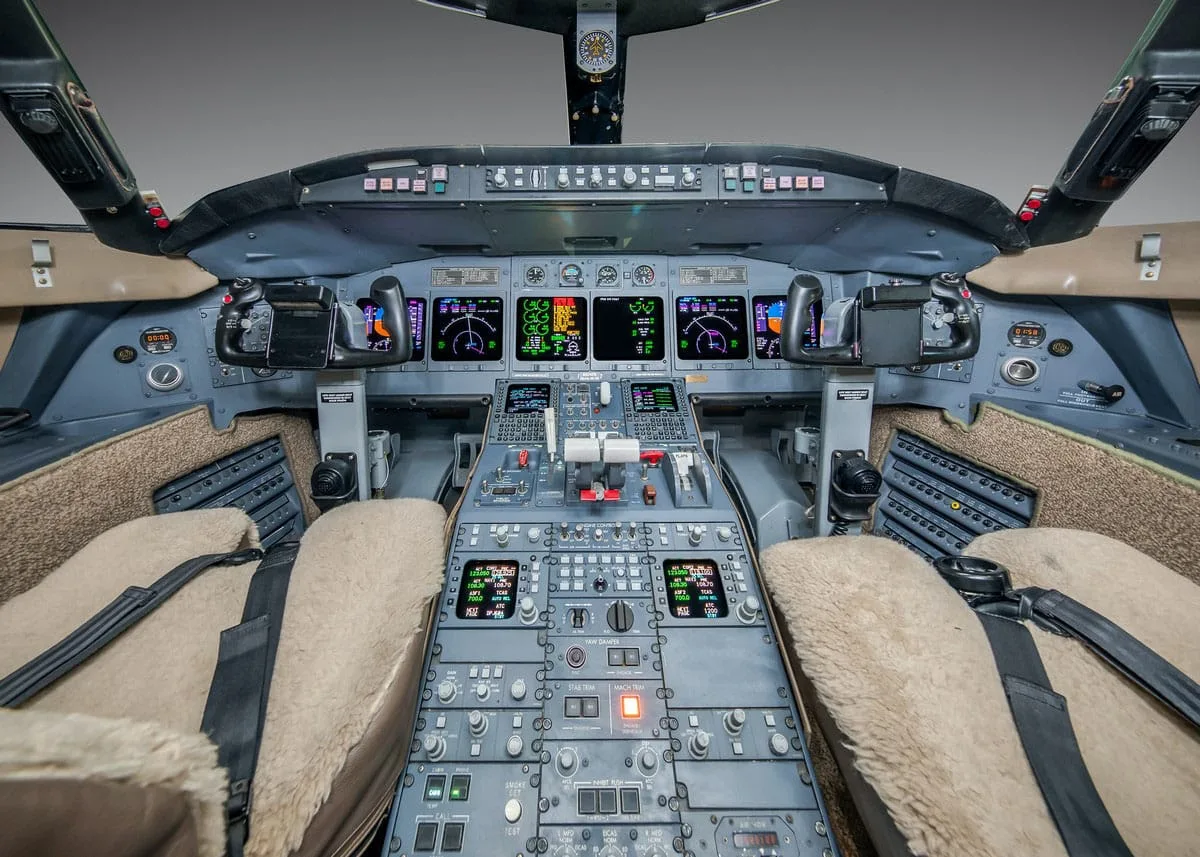 The cockpit of a business jet, showing multiple digital displays, control yokes, and a central control panel with numerous buttons and switches.