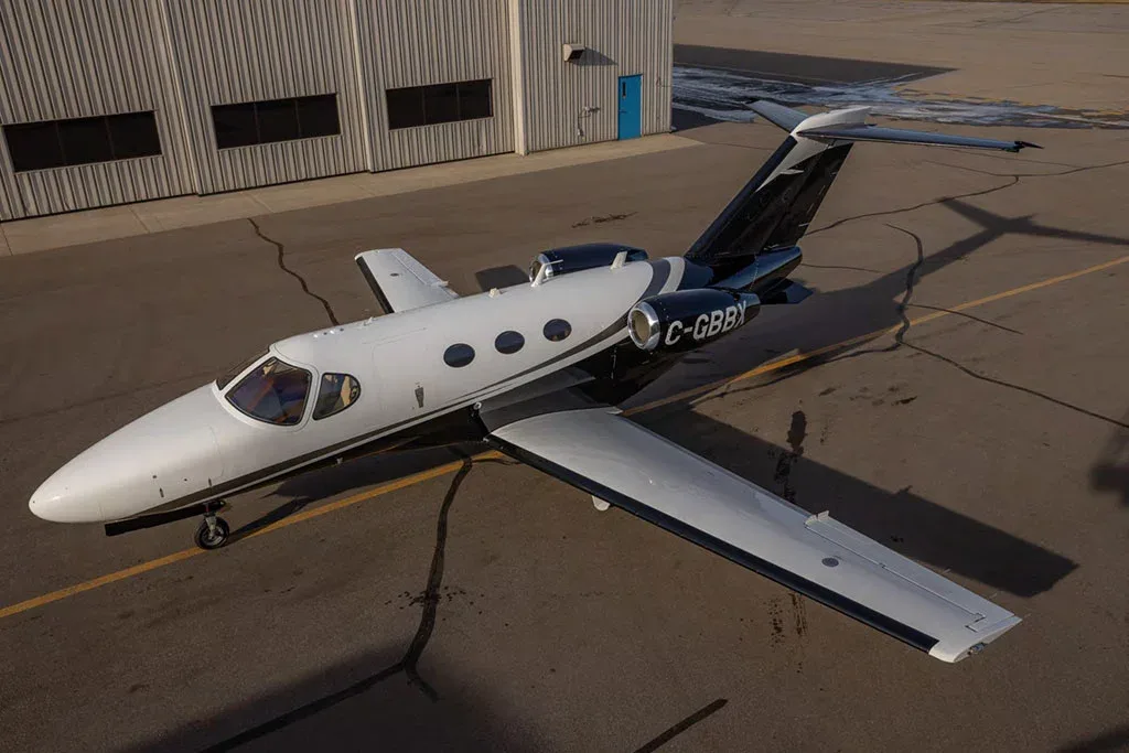 An aerial view of a white and black private jet, registration C-GBBK, parked on an asphalt apron next to a hangar.