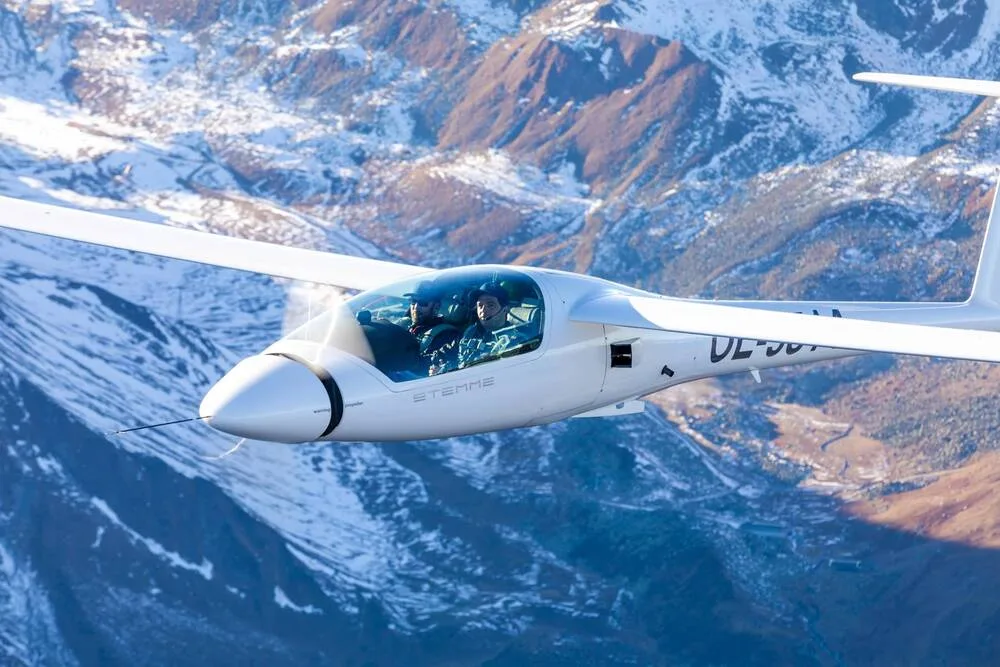 A white Stemme motorglider with two pilots in the cockpit flying over snowy mountains.