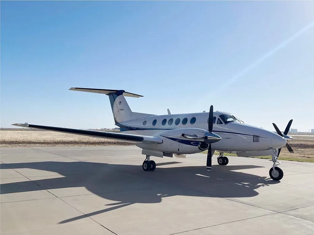 A white and blue twin-turboprop aircraft, likely a King Air, is parked on an airfield under a clear blue sky.