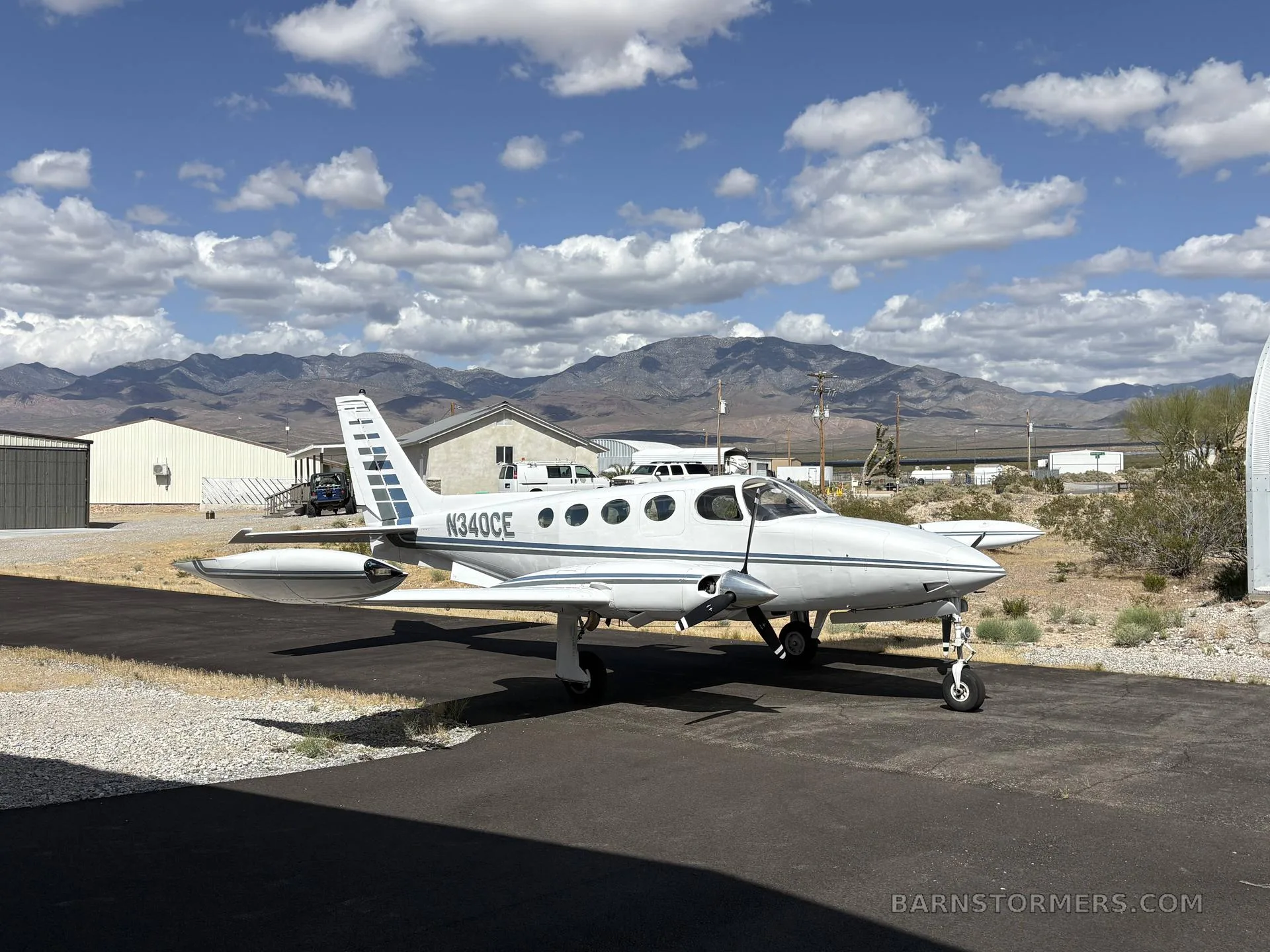 A white Cessna 340 (N340CE) with blue stripes parked on an asphalt ramp with mountains in the background.
