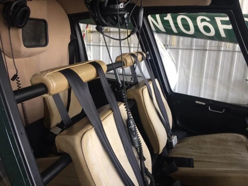 The tan leather seats and seatbelts of a helicopter cabin are visible, with headsets hanging nearby.