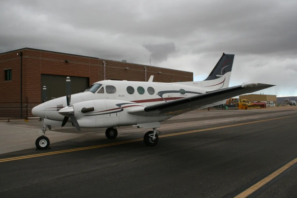 A side view of a white twin-engine turboprop aircraft with dark red and blue stripes, parked on an asphalt runway next to a brick building.