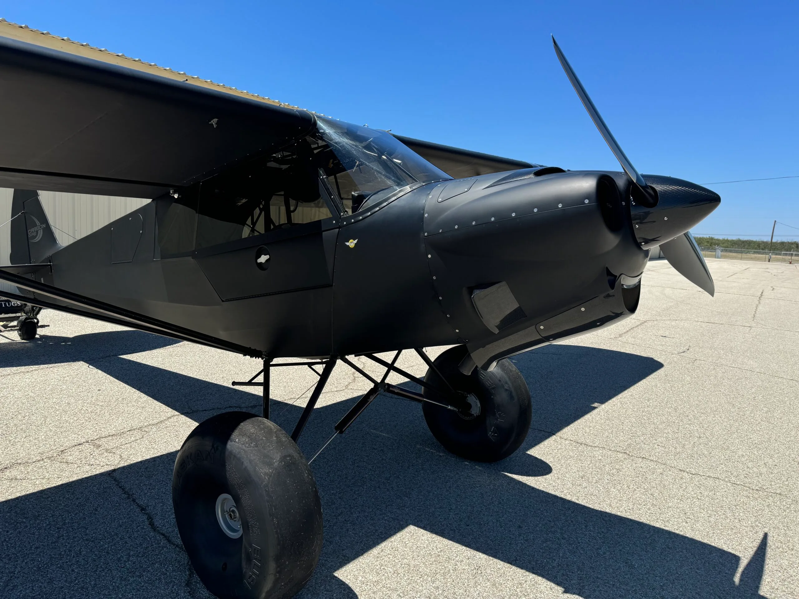 A black Carbon Cub-style aircraft with large bush tires is parked on an asphalt apron under a clear blue sky.