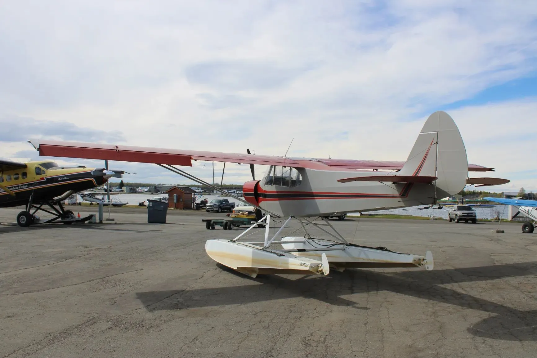 A rear-side view of a red and grey floatplane with N984 on the tail, parked on an apron with other aircraft and buildings.