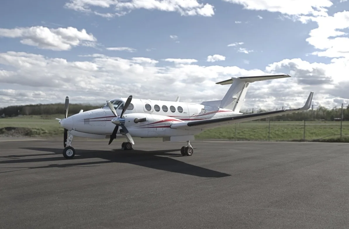 A white twin-turboprop aircraft with red accent stripes is parked on a tarmac under a partly cloudy sky.