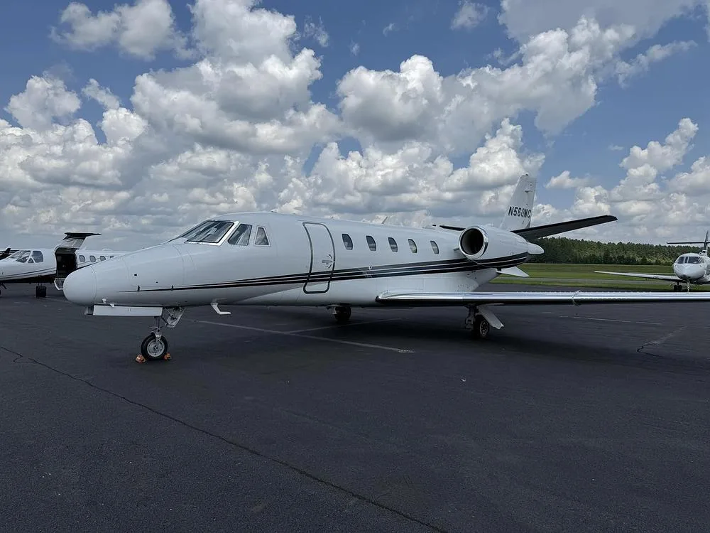 A white Cessna Citation private jet with black and grey stripes parked on an airport tarmac with other aircraft in the background.