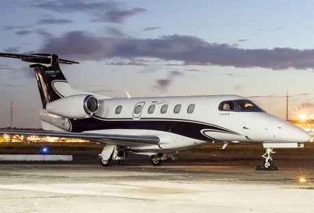 A Phenom 300E aircraft with a black and white paint scheme is parked on the tarmac at dusk.