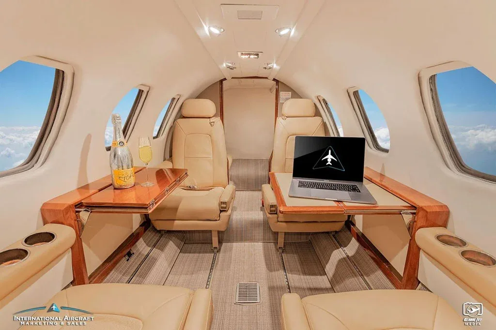 The luxurious cabin interior of a business jet with beige leather seats, wooden tables, and a laptop on one table.