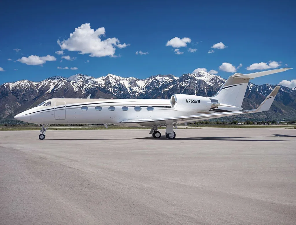 A side view of a white Gulfstream G650 business jet with registration N769MM on a tarmac, with mountains under a blue sky.