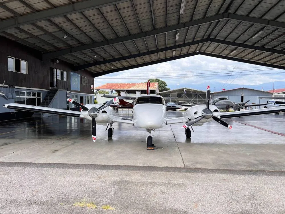 A white twin-engine propeller aircraft with red stripes on its propellers, parked head-on under a hangar.