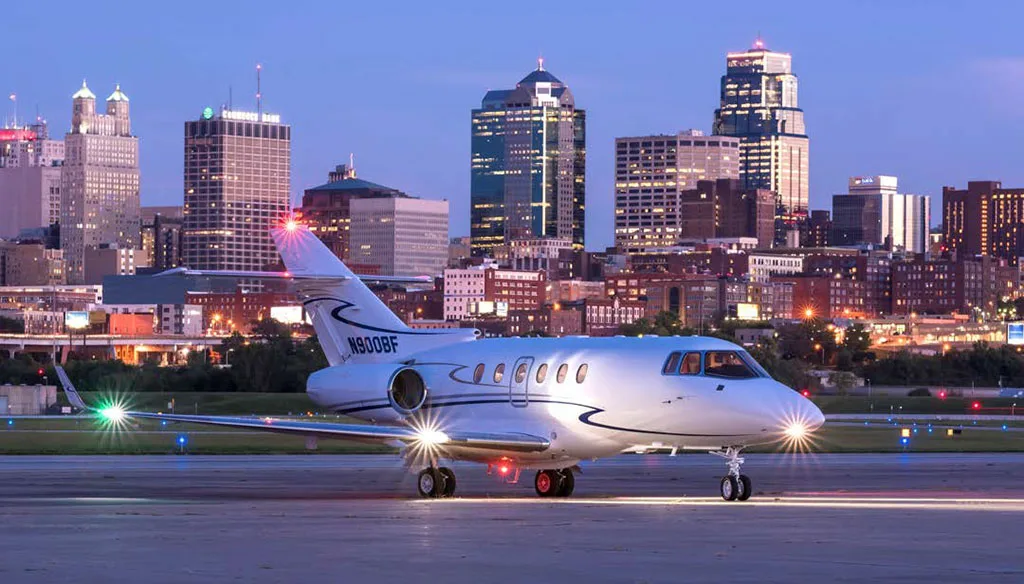 A white Hawker 900XP business jet with registration N900BF is parked on a tarmac at dusk, with the illuminated Kansas City skyline in the background.