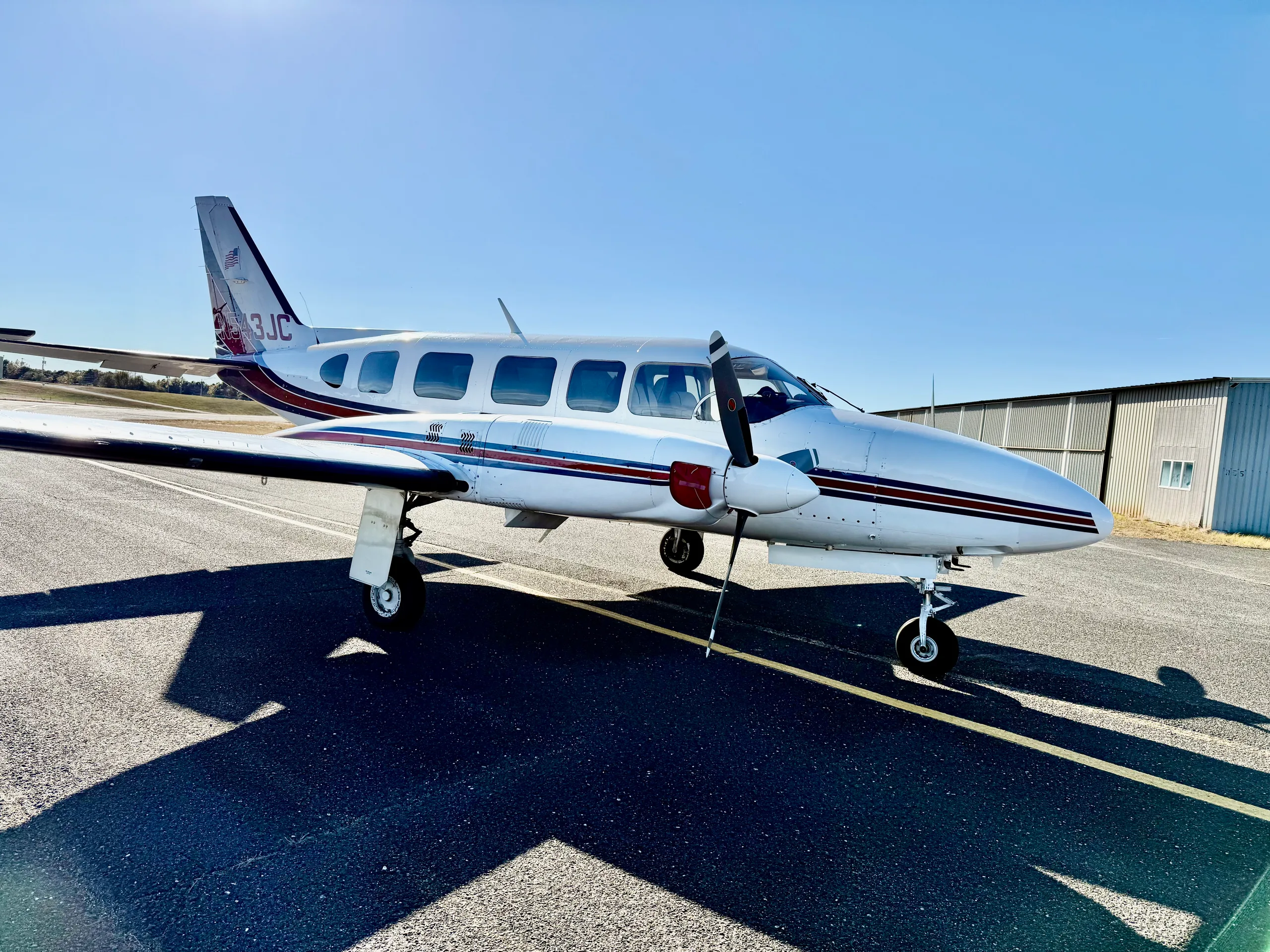 A side view of a white twin-engine propeller aircraft with red and blue stripes and registration N543JC on the tail, parked on an asphalt ramp.