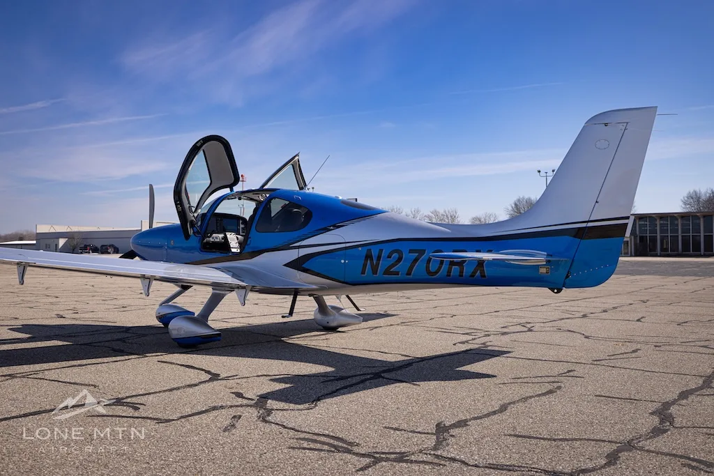 A rear-side view of the blue and silver Cirrus SR20 aircraft, registration N270RX, on an airport tarmac with its doors open.