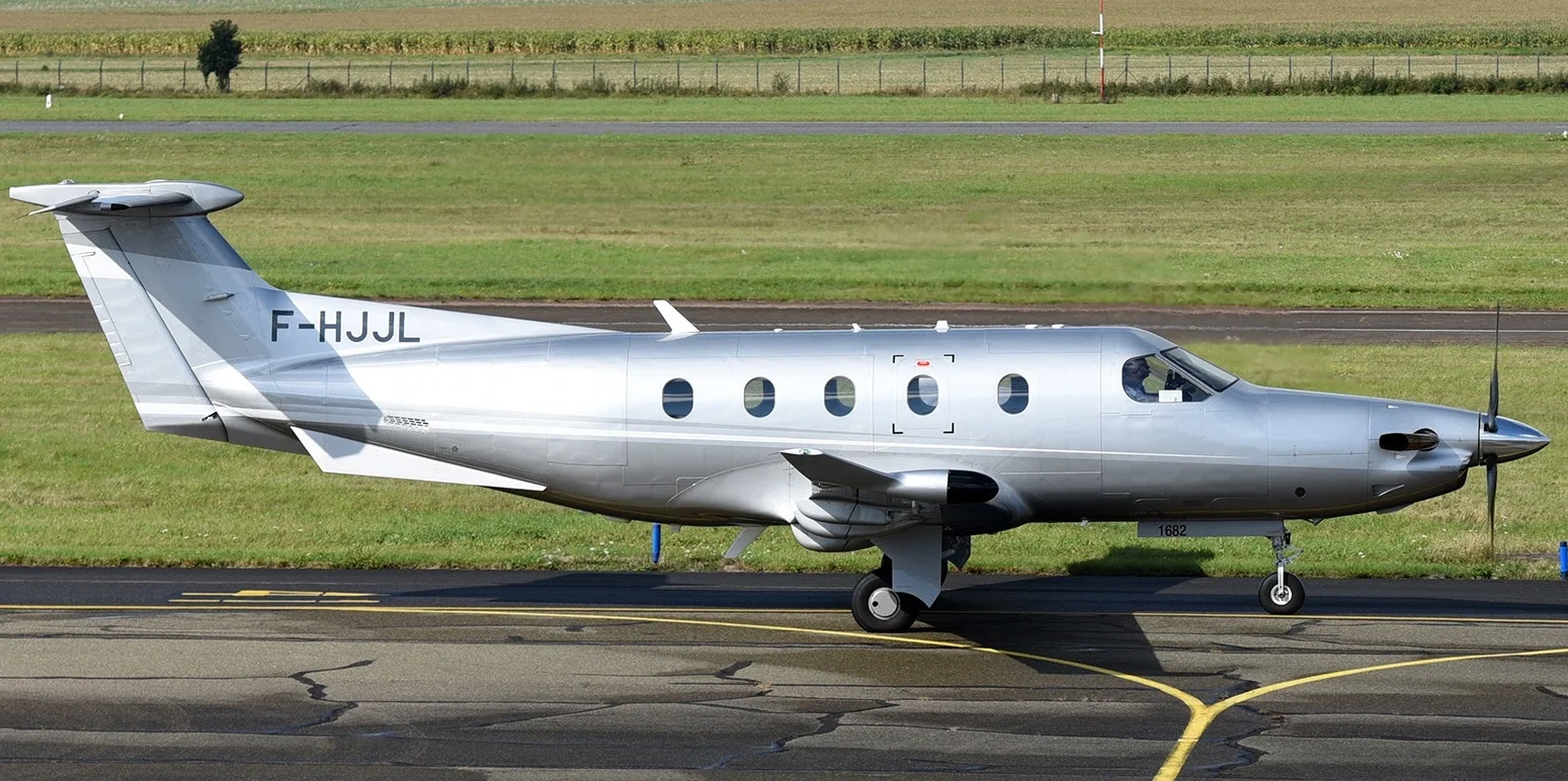 A silver Pilatus PC-12 aircraft with registration F-HJJL taxiing on an airfield, viewed from the side with green fields in the background.