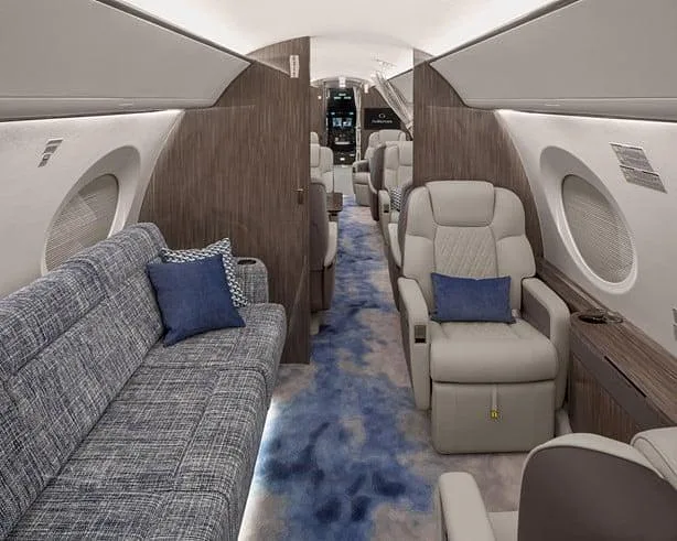 A private jet's interior cabin features a grey fabric sofa on the left and a single light grey quilted seat on the right, with a blue patterned carpet.