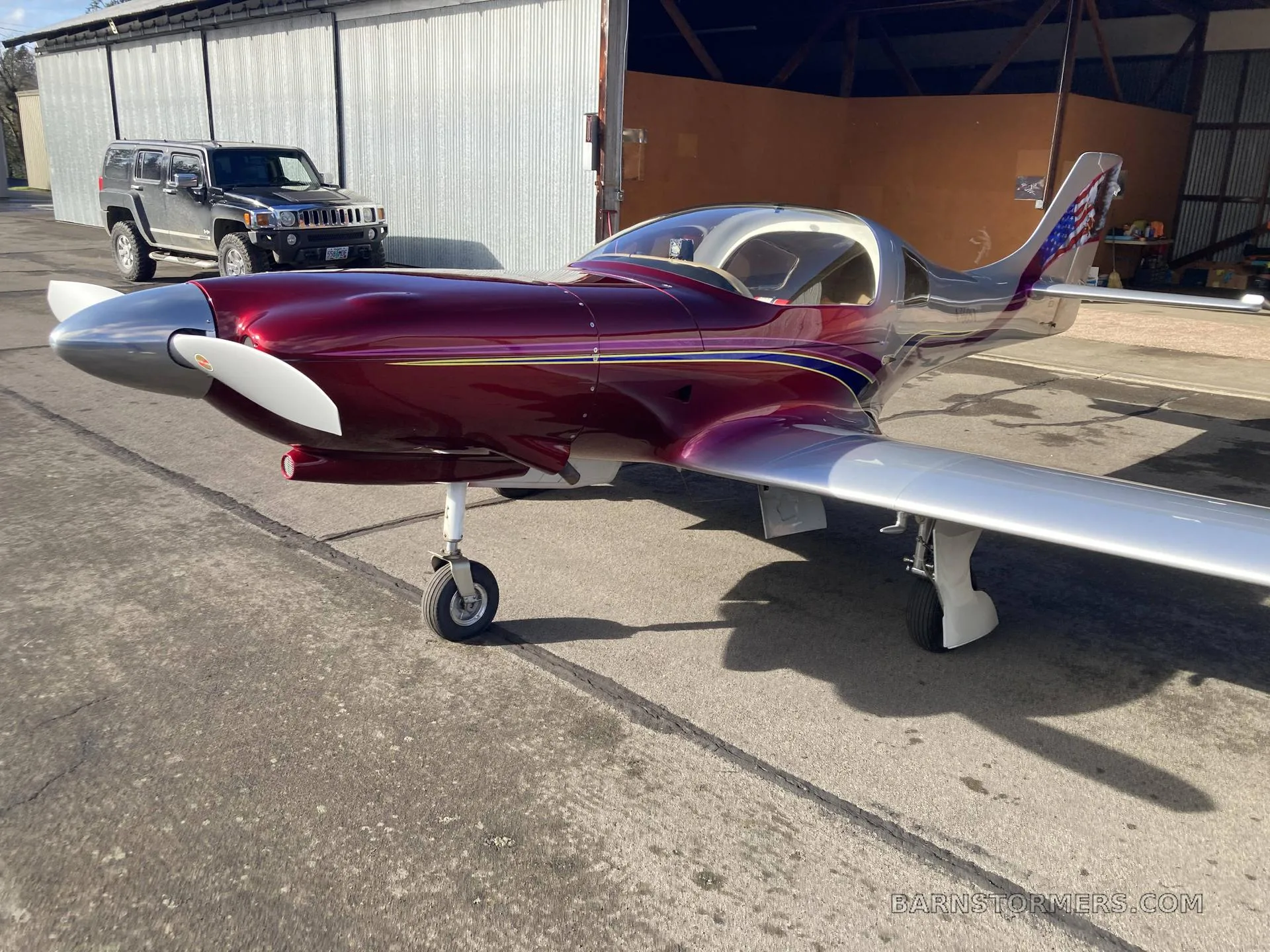 A metallic red and silver Lancair 360 aircraft with a white propeller, parked on an asphalt ramp in front of a hangar with a black SUV nearby.