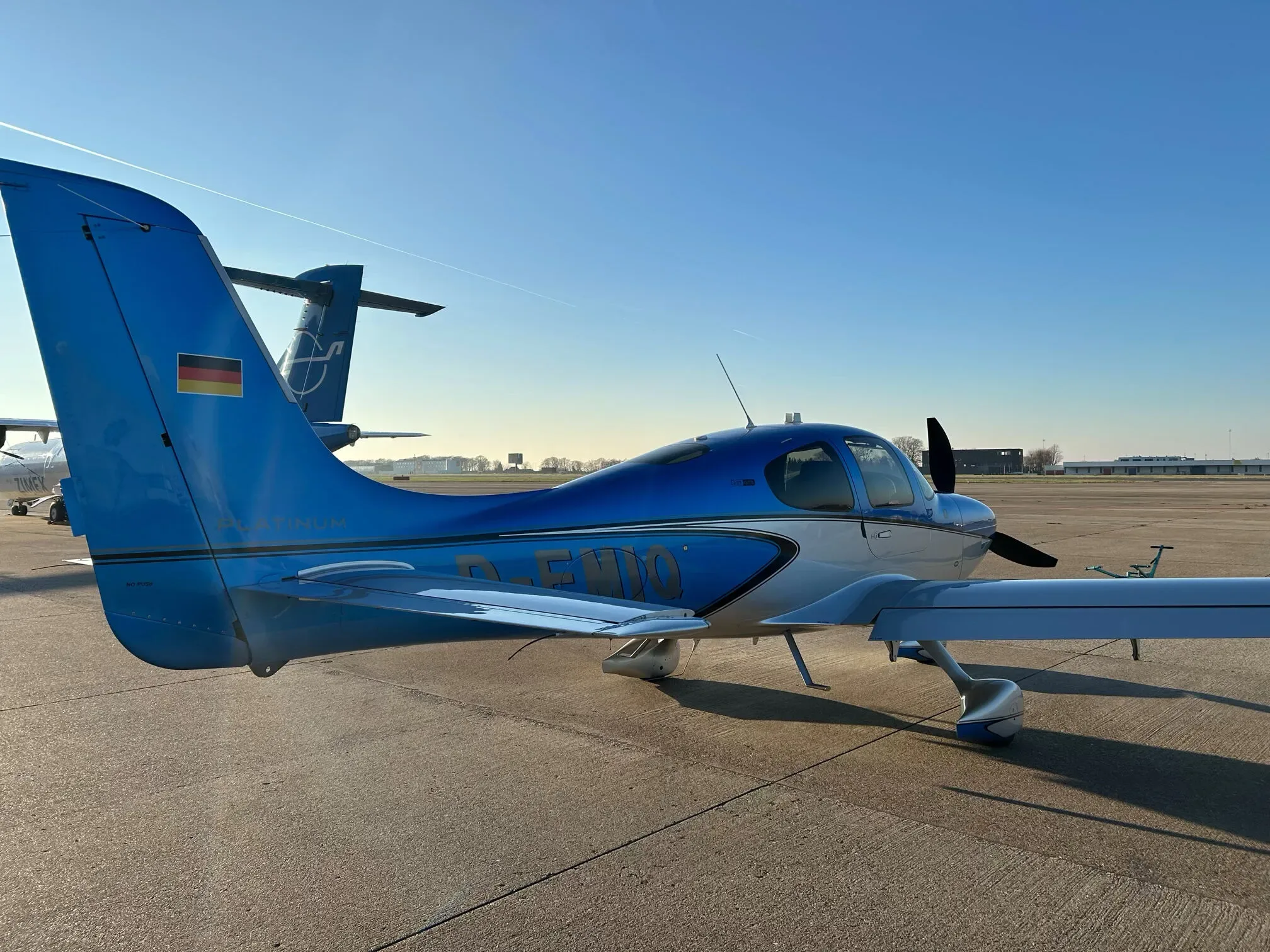 A rear-side view of a blue and silver Cirrus SR22 aircraft with the German flag on its tail, parked on an airfield.