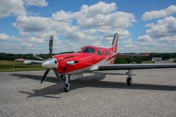 A red and white Piper M600 SLS aircraft with registration N325ST is parked on an asphalt ramp under a partly cloudy sky, viewed from the front-left.